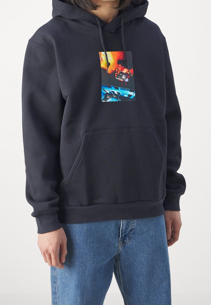 Person wearing a black hoodie with front pocket and a colorful graphic showing a person holding shoes, paired with blue jeans.