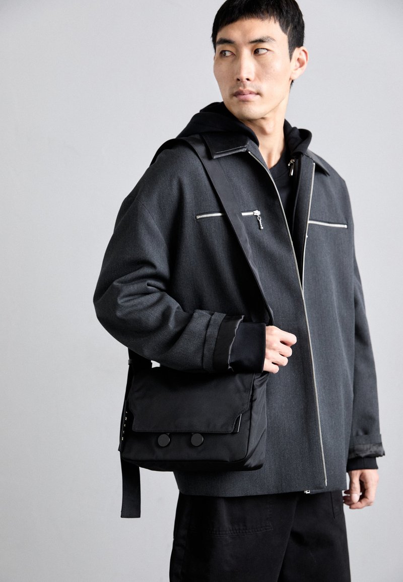 Sort skuldertaske lavet af nylon, med en klap forsynet med to knappelukninger. Båret med en tekstureret grå jakke med lynlås-lommer.