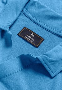 Mėlynas audinio polo marškinėliai su minkštu paviršiumi. Savybė - juodas ženklas su auksinėmis siūlėmis, kuriame užrašyta "Martinique Heritage Apparel."