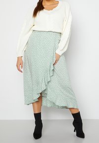 Vrouw in een wit kabelbreiselsweater, groene bloemen midi-jurk met gerimpelde zoom en zwarte enkellaarsjes met hak, staand tegen een effen muur.