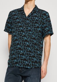 Chemise noire à manches courtes présentant un motif abstrait de texte bleu, confectionnée dans un tissu léger avec un col cranté et une fermeture boutonnée à l'avant.