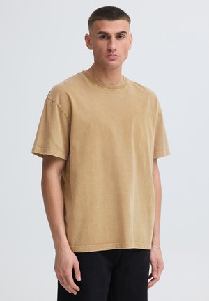 Jeune homme aux cheveux courts portant un t-shirt ample beige et un pantalon noir, debout devant un fond clair uni.