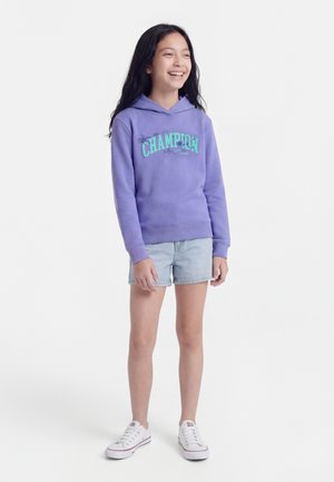 Young girl smiling, wearing a purple hoodie with "CHAMPION" text, light denim shorts, and white sneakers, standing on a plain white background.