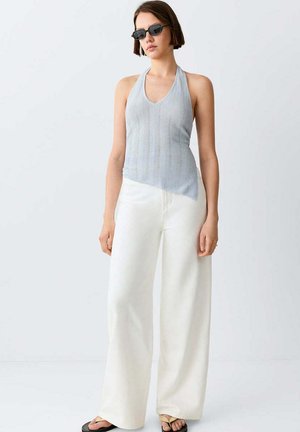 Mujer con gafas de sol, una blusa sin mangas halter de color gris claro, pantalones blancos de pierna ancha y sandalias beige sobre un fondo claro liso.