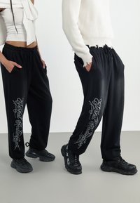 Black sweatpants with side pockets, featuring silver script detail along the outer leg. Made from a soft, textured fabric.