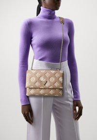 Sac à main en cuir beige avec motif géométrique blanc, bandoulière en chaîne et détail de logo triangulaire. Tenue par une personne portant un col roulé violet.