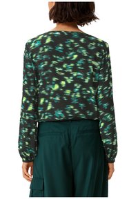 Gekürzte schwarze Bluse mit grünem und türkisfarbenem abstraktem Muster, langen Ärmeln und elastischen Manschetten, kombiniert mit hoch taillierten dunkelgrünen Hosen.