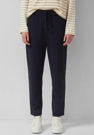 Personne portant un pantalon noir à cordon, un t-shirt à manches longues rayé beige et blanc, et des baskets blanches, debout devant un fond gris uni.