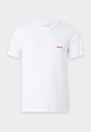Mann steht barfuß, trägt ein weißes Kurzarm-T-Shirt mit kleinem roten Text auf der Brust und schwarze enganliegende Shorts vor einfarbigem Hintergrund.