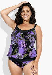 Femme souriante aux cheveux courts et foncés portant un maillot tankini violet à motifs floraux noirs, une main sur la tête, sur fond blanc.