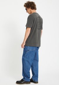 Maglietta grigia in cotone con una grande grafica sul retro, abbinata a pantaloni di denim blu e scarpe nere stringate.