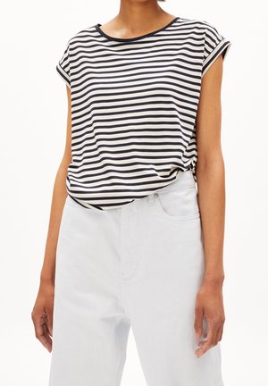 Frau trägt ein schwarz-weiß gestreiftes T-Shirt mit kurzen Ärmeln, das in eine hoch taillierte weiße Hose gesteckt ist, vor schlichtem Hintergrund.