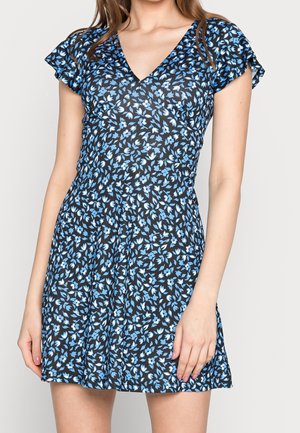 Femme portant une robe noire courte avec un motif floral bleu et blanc et des manches flottantes.