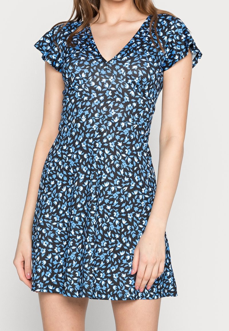 Femme portant une robe noire courte avec un motif floral bleu et blanc et des manches flottantes.