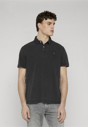 Ung mann med kort krøllete hår iført en falmet svart poloskjorte og lysegrå jeans, ser mot høyre mot en ensfarget bakgrunn.