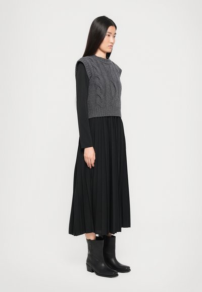 Pelēka adīta cropped džemperis virs melnas garām piedurknēm kleitas ar plisētu svārku, apvienots ar melniem potīšu zābakiem. Modelis stāv šķērsām.
