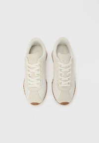 Μπεζ σουέντ αθλητικά παπούτσια με λευκά κορδόνια, υφασματική λεπτομέρεια στο πλάι και σόλες από καουτσούκ σε χρώμα τσίχλας. Διαθέτουν επενδεδυμένο γιακά και μινιμαλιστικό σχεδιασμό.