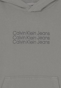 Felpa grigia chiara con tasca frontale e testo ripetuto "Calvin Klein Jeans" ricamato in scuro blu navy. Texture del tessuto liscia.