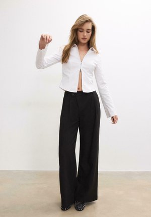 Femme debout, yeux fermés, portant un haut court blanc à fermeture éclair, un pantalon noir à jambes larges et des chaussures noires cloutées sur un fond neutre.