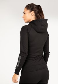 Zwarte hoodie met een slank ontwerp, met een capuchon en geribbelde manchetten. De linker mouw toont "GORILLWEAR" in witte letters.