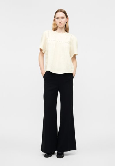 Γυναίκα στέκεται με τα χέρια στις τσέπες, φορώντας ένα εκρού μπλουζάκι με κοντά μανίκια και ψηλόμεσα μαύρα παντελόνια φαρδιάς γραμμής με μαύρα παπούτσια.