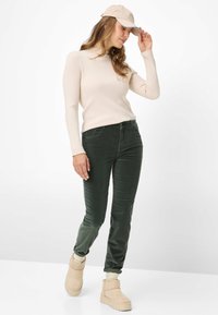 Beige ribbet langærmet top, grønne fløjlsbukser, tanfarvede ankelstøvler og en lys kasket. Glat tekstur med tætsiddende pasform.