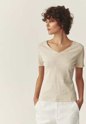 Kvinde med krøllet hår iført en beige V-hals kortærmet trøje og hvide bukser, som kigger mod højre med hænderne i lommerne.