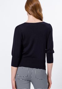 Marineblauwe gebreide trui met driekwart pofmouwen, geribbelde zoom en een ronde halslijn. Gepaarde met zwart-wit geruite broeken.