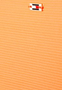 Tecido laranja claro com uma superfície texturizada e em nervuras. Apresenta um emblema retangular com um design em preto, branco e vermelho.