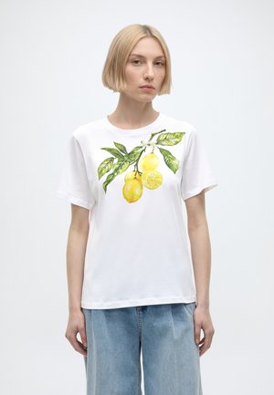 Femme blonde portant un t-shirt blanc avec un imprimé branche de citron et un jean large bleu clair sur fond uni.