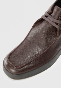Zapato casual de cuero marrón con un acabado texturizado, punta redondeada y suela de goma negra de contraste. Cuenta con un cierre de cordones delgados y acentos de costura.