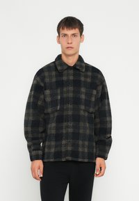 Jachetă din fleece cu carouri în nuanțe de bleumarin și bej, dotată cu guler, două buzunare pe piept și închidere cu butoane. Textură moale și croială lejeră.
