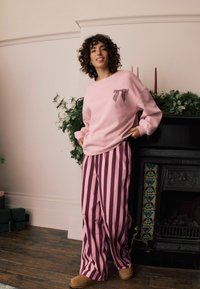 Jasnoróżowy sweter z czarno-białą, paskowaną kieszenią w kształcie kokardy, w zestawieniu z różowymi i ciemnoróżowymi, paskowanymi spodniami o szerokich nogawkach oraz brązowymi klapkami.