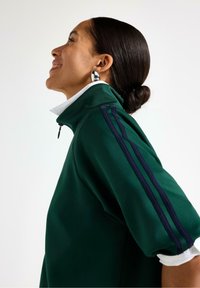 Grøn sweatshirt med høj krave og hvid krave, med marineblå sidelister og halv lynlås, vist på en model, der står med siden til.