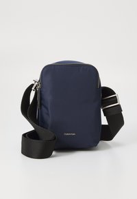 Borsa a tracolla in nylon blu marina con chiusura a zip, tracolla nera regolabile, hardware argentato e logo Calvin Klein sul davanti.