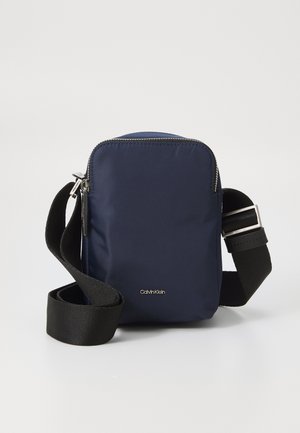 Geantă de umăr din nylon de culoare navy, cu închidere cu fermoar, curea ajustabilă neagră, elemente de metal argintii și logo Calvin Klein pe față.