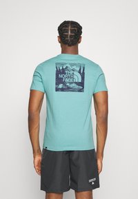 Tyrkysové bavlněné tričko s tmavomodrou grafikou horských a stromových motivů, logo značky na zadní straně, v kombinaci s tmavými kraťasy.