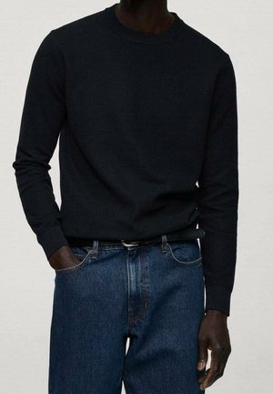 Uomo che indossa un maglione nero aderente a maniche lunghe infilato in jeans di denim blu con una cintura nera, con una mano in tasca.