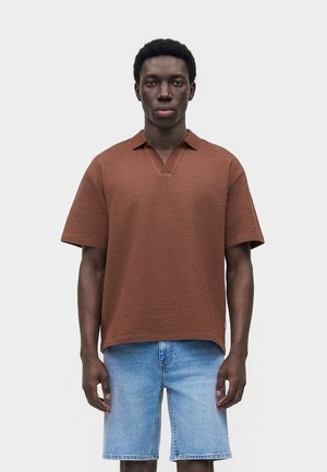 Jeune homme portant une chemise marron texturée à manches courtes et un short en jean bleu clair, debout devant un fond blanc uni.