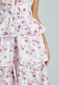 Weißes Blumenkleid mit Peplum-Design, Rüschenschichten, rosa gestickten Blumen, Lochmuster-Details und einem runden Stoffgürtelakzent.