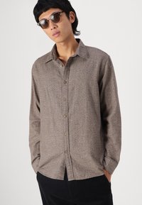 Καφέ πουκάμισο με σχέδιο, με κολάρο, μακριά μανίκια και άνετη εφαρμογή, συνδυασμένο με σκούρα παντελόνια και γυαλιά ηλίου.