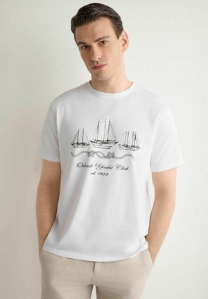 Młody mężczyzna w białej koszulce z czarnymi szkicami trzech jachtów i napisem "Ochnik Yacht Club est. 1989".