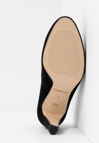 Chaussure à talon haut en daim noir avec une semelle en caoutchouc beige lisse. Présente des motifs ondulés subtils et un logo imprimé à l'intérieur de la semelle.