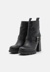 Botines negros de tobillo hechos de cuero sintético texturizado, con un tacón grueso, correa con detalle de hebilla metálica y lengüetas para ponerlos y quitarlos fácilmente.