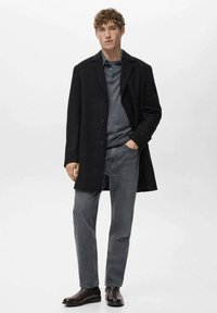 Abrigo de lana negro con un corte entallado, diseño de un solo botón, llevado sobre un suéter gris oscuro y jeans grises, complementado con botas de cuero negras.