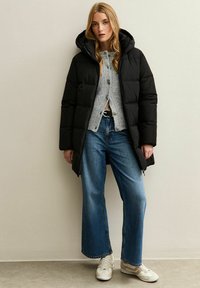 Cappotto imbottito nero con cappuccio, caratterizzato da un design trapuntato, indossato sopra un maglione grigio e abbinato a jeans a gamba larga blu e sneakers beige.