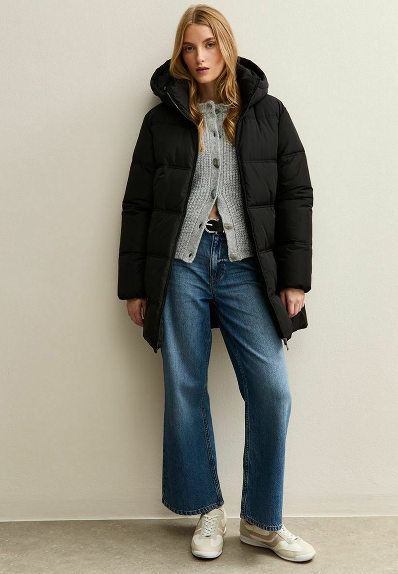 Cappotto imbottito nero con cappuccio, caratterizzato da un design trapuntato, indossato sopra un maglione grigio e abbinato a jeans a gamba larga blu e sneakers beige.