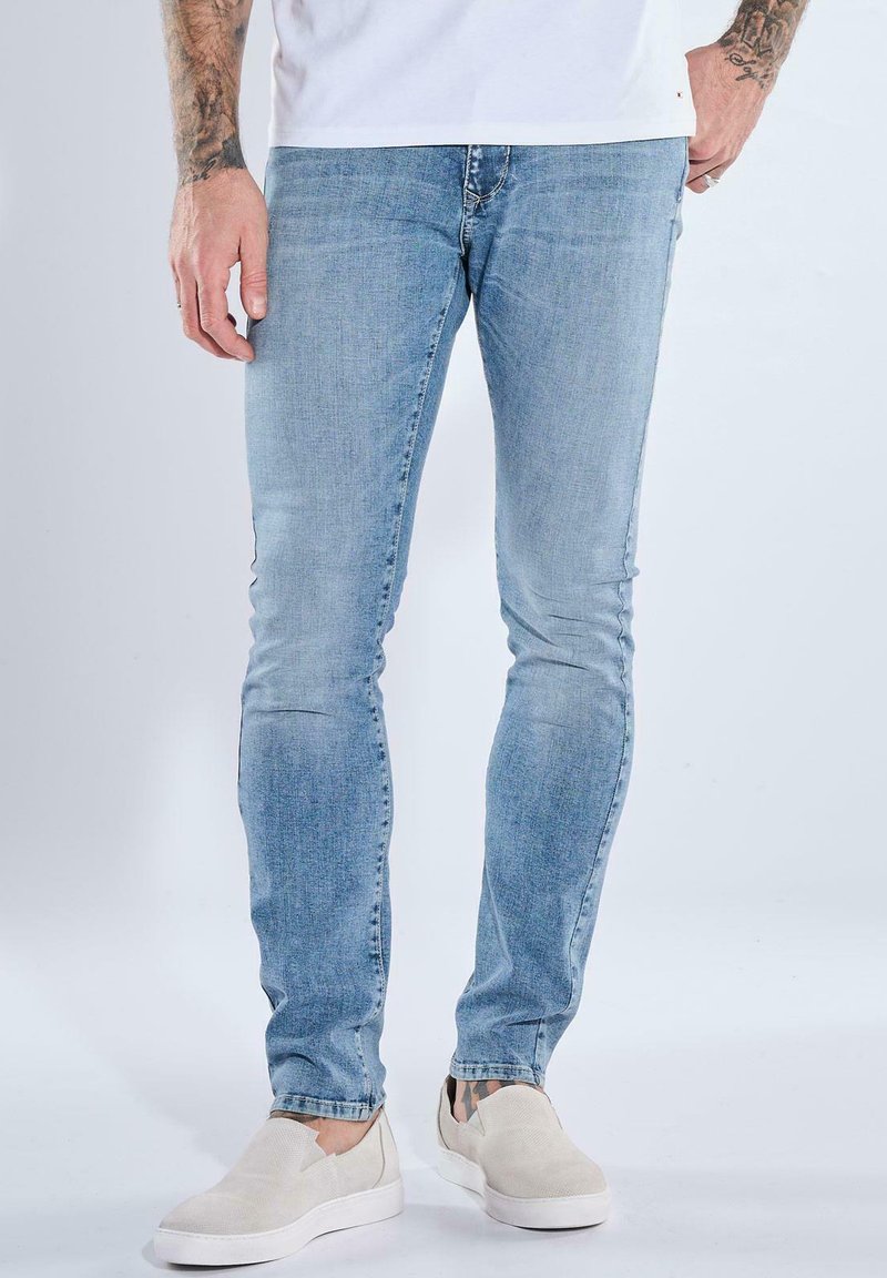 Man wearing light blue slim-fit jeans and beige slip-on shoes, standing with hands relaxed by sides on a white background.