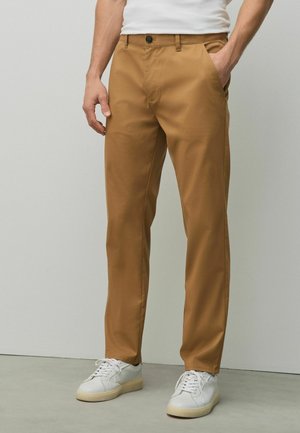 Mann trägt beige gerade geschnittene Hose mit Seitentaschen und weißen Sneakers, steht vor einem einfarbigen hellgrauen Hintergrund.