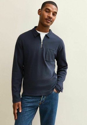 Polo a maniche lunghe blu navy con colletto a mezza zip, caratterizzato da una tasca sul petto e una texture liscia, abbinato a jeans blu.